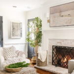 Cozy neutral living room with a white chair, plants, lit, limewashed brick fireplace and snow art on the wall.