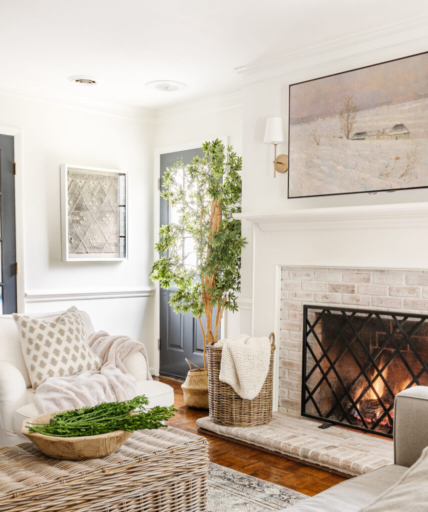 Cozy neutral living room with a white chair, plants, lit, limewashed brick fireplace and snow art on the wall.